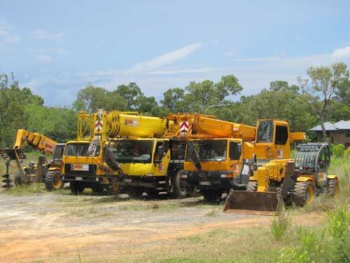 A Row of Yellow Construction Vehicles Are Parked In A Dirt Field — Smithys Crane Hire in Trinity Beach, QLD