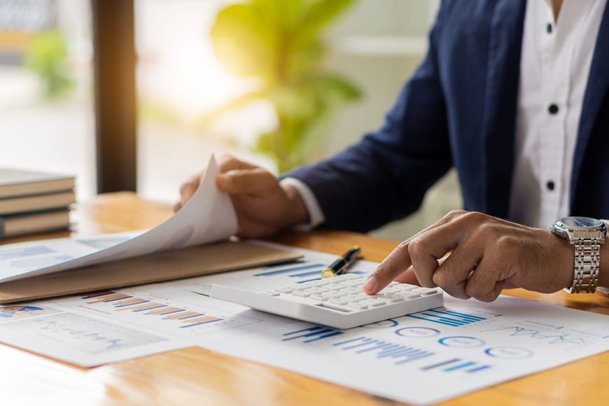 Person in a blue blazer using a calculator, reviewing papers with financial charts at a desk.