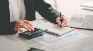 Person using a calculator and pen, working with financial documents at a desk near a laptop.