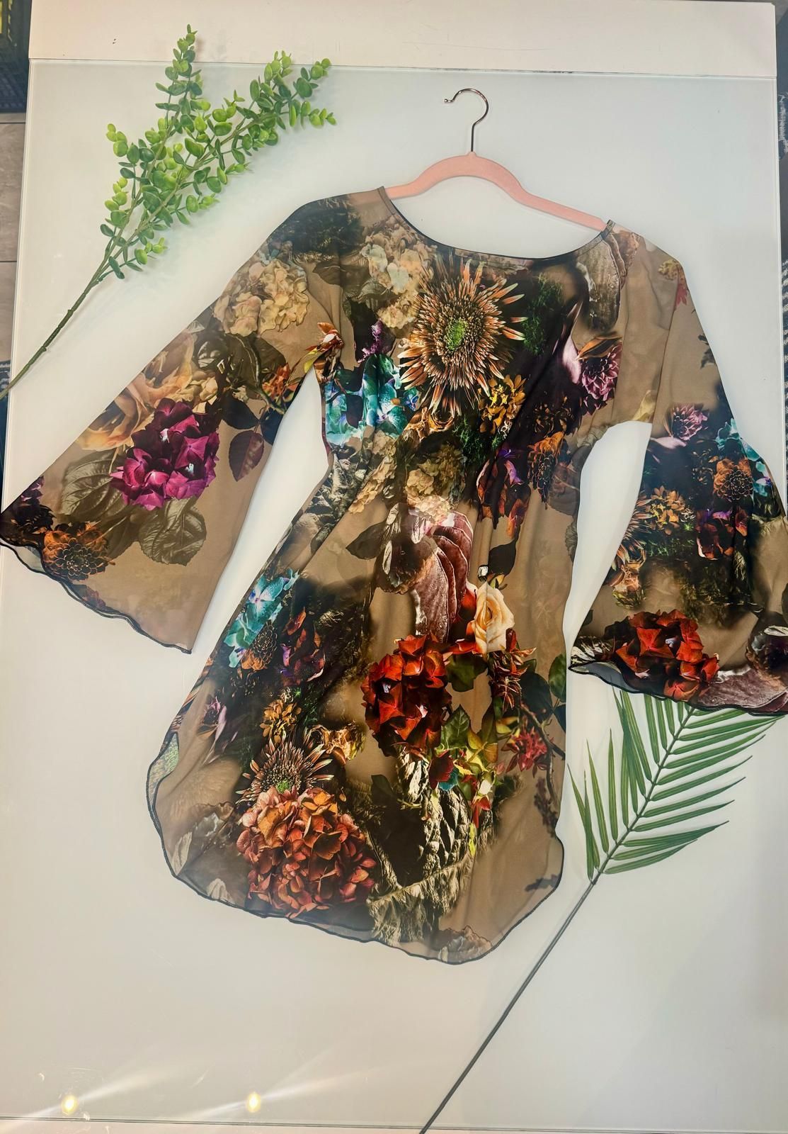 Un abito floreale con maniche a campana è appeso a una gruccia accanto a una pianta.