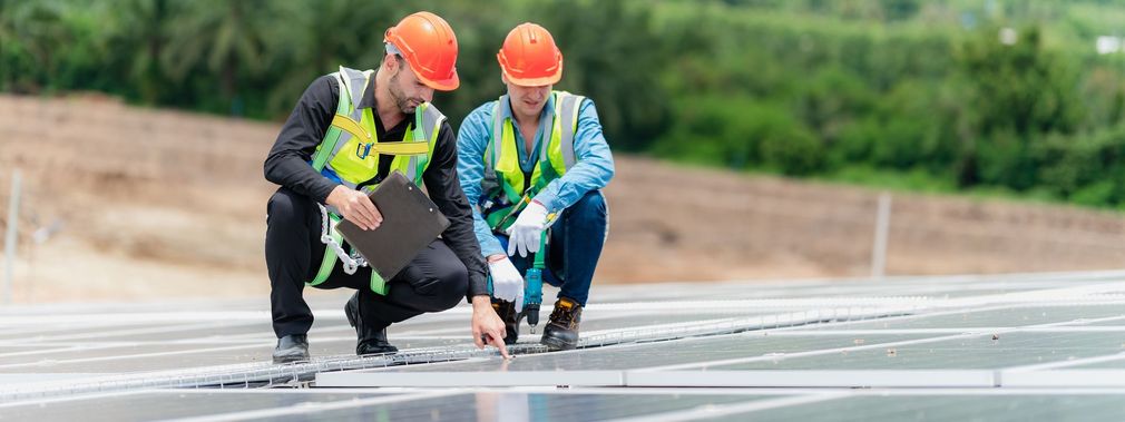 Two construction workers are kneeling down and looking at a solar panel.