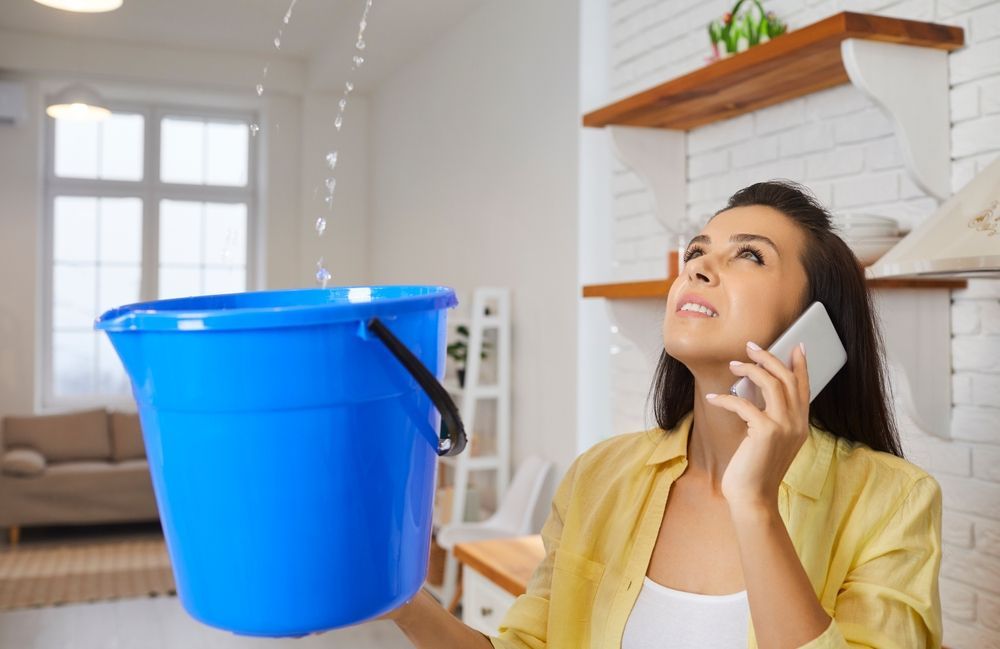 Woman on phone watches water leak into blue bucket in home.