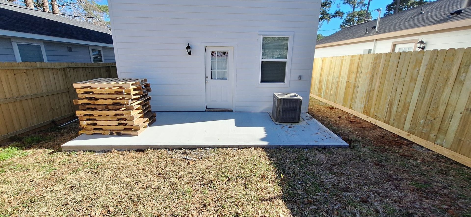 A white house with a wooden fence and a stack of wooden pallets in the backyard.