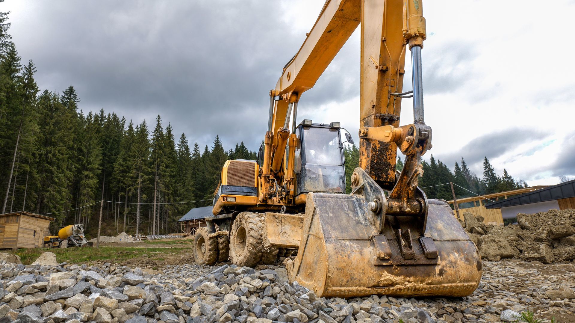 Yellow excavator digging rocks at a construction site under cloudy skies, surrounded by pine trees.