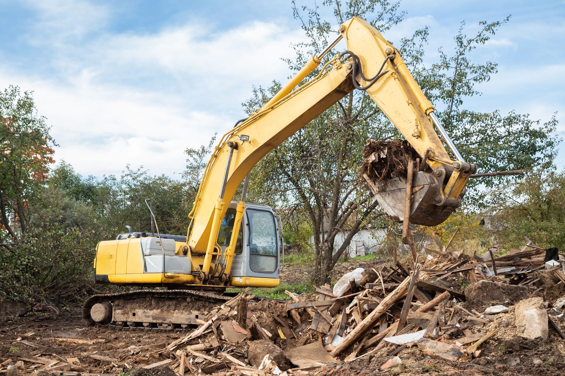 Yellow excavator clearing debris in a wooded area under a cloudy sky