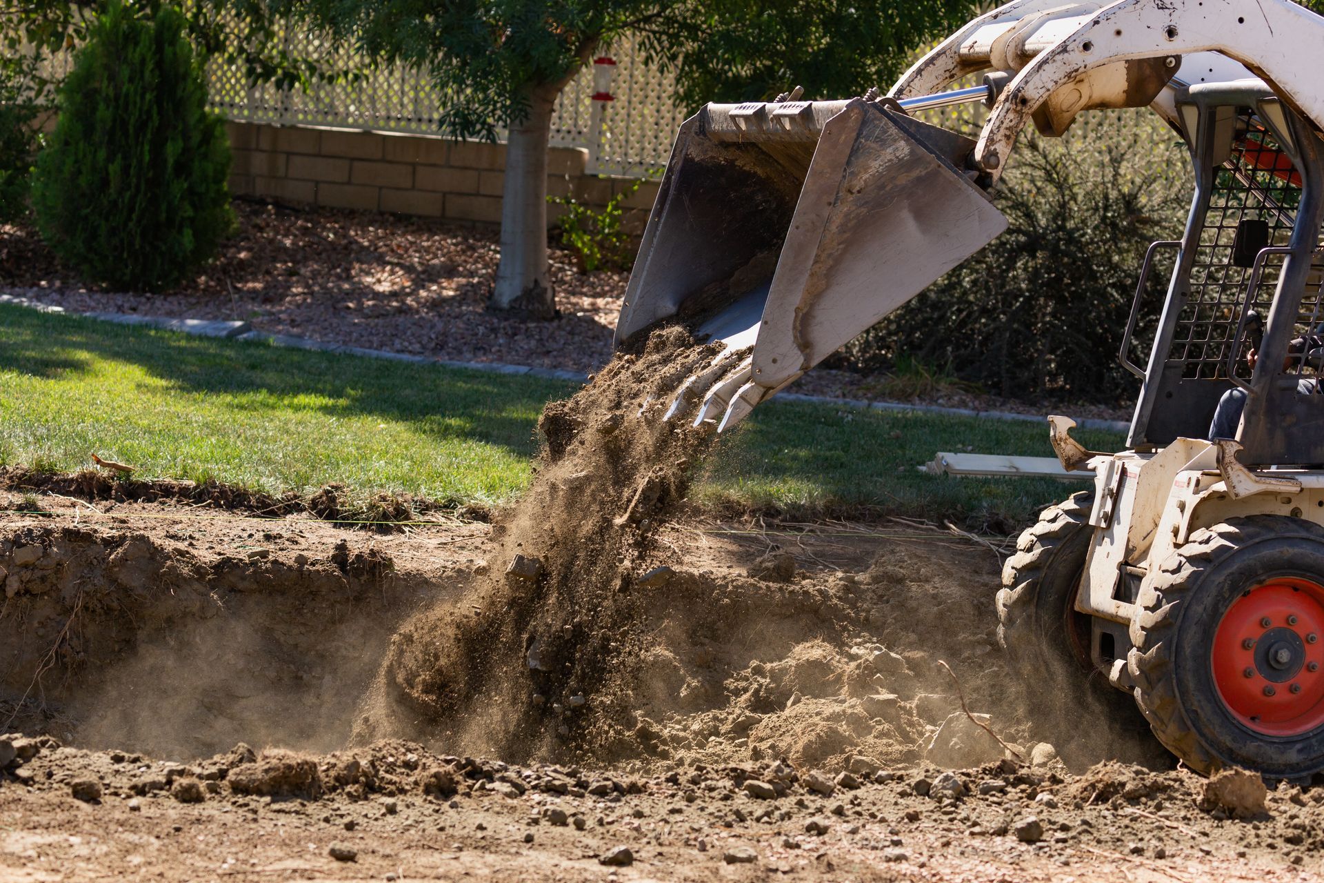 A construction loader dumping soil into a trench in a yard with trees and a stone wall. A construction loader dumping soil into a trench in a yard with trees and a stone wall.