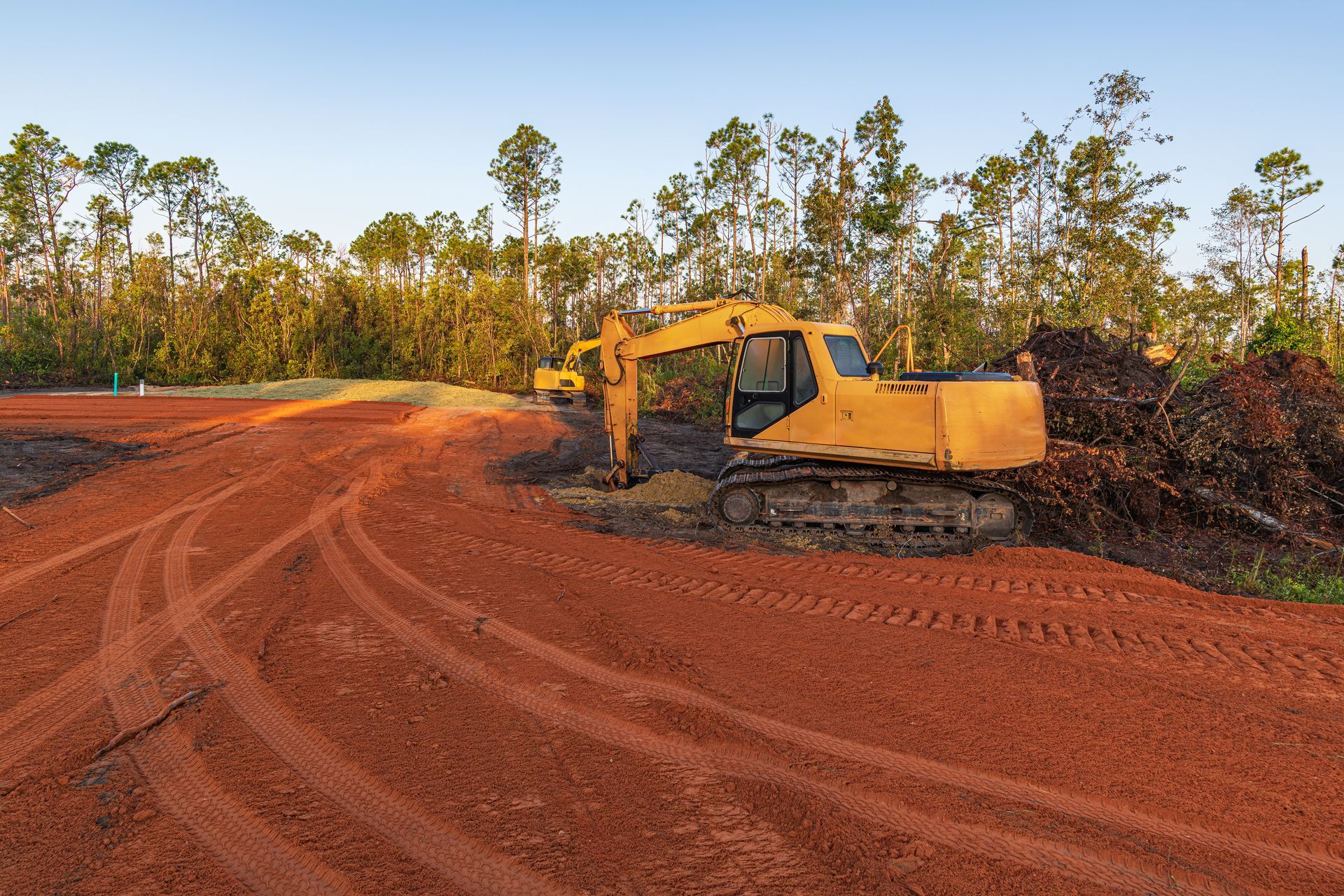 Yellow excavator on red dirt at a cleared construction site with trees in the background