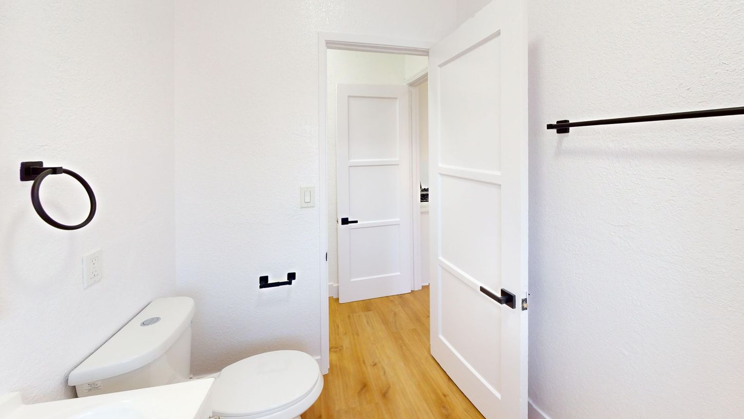 Bathroom with white walls, wooden floor, toilet, black towel bar and door handles.