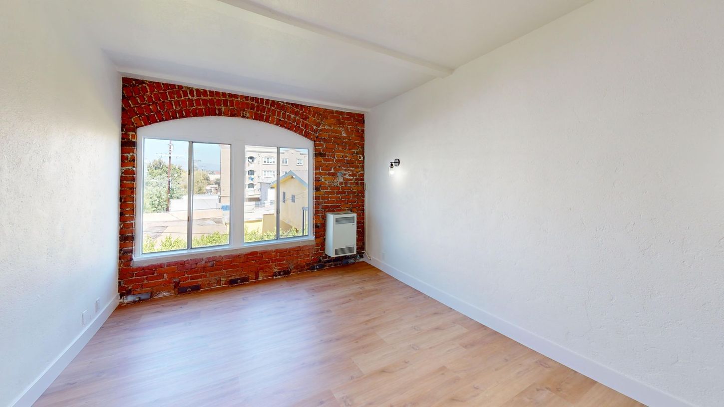 Empty room with brick accent wall and large arched window. Wooden floor, white walls.