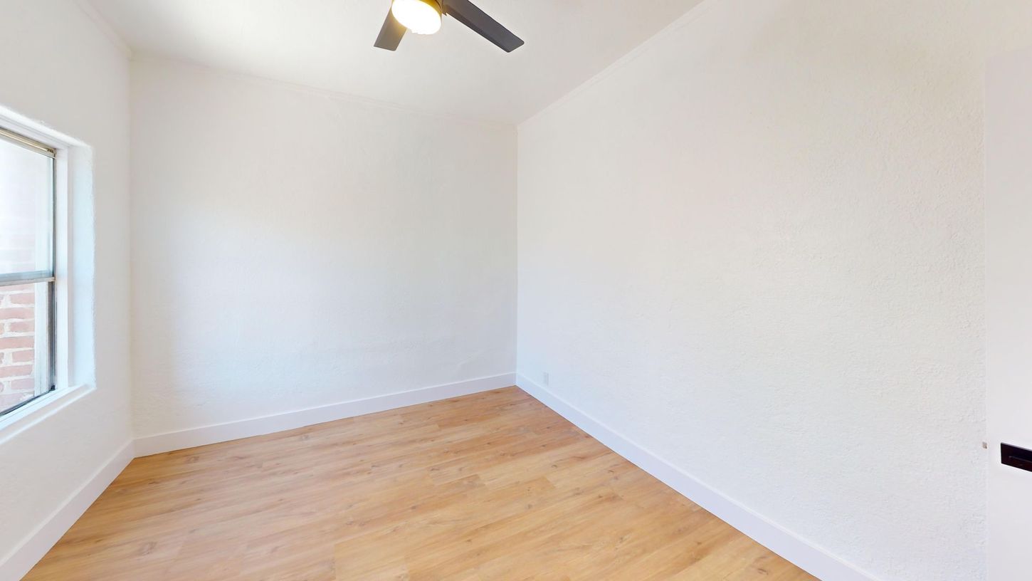 Empty room with hardwood floors, white walls, and a ceiling fan. A window is on the left.