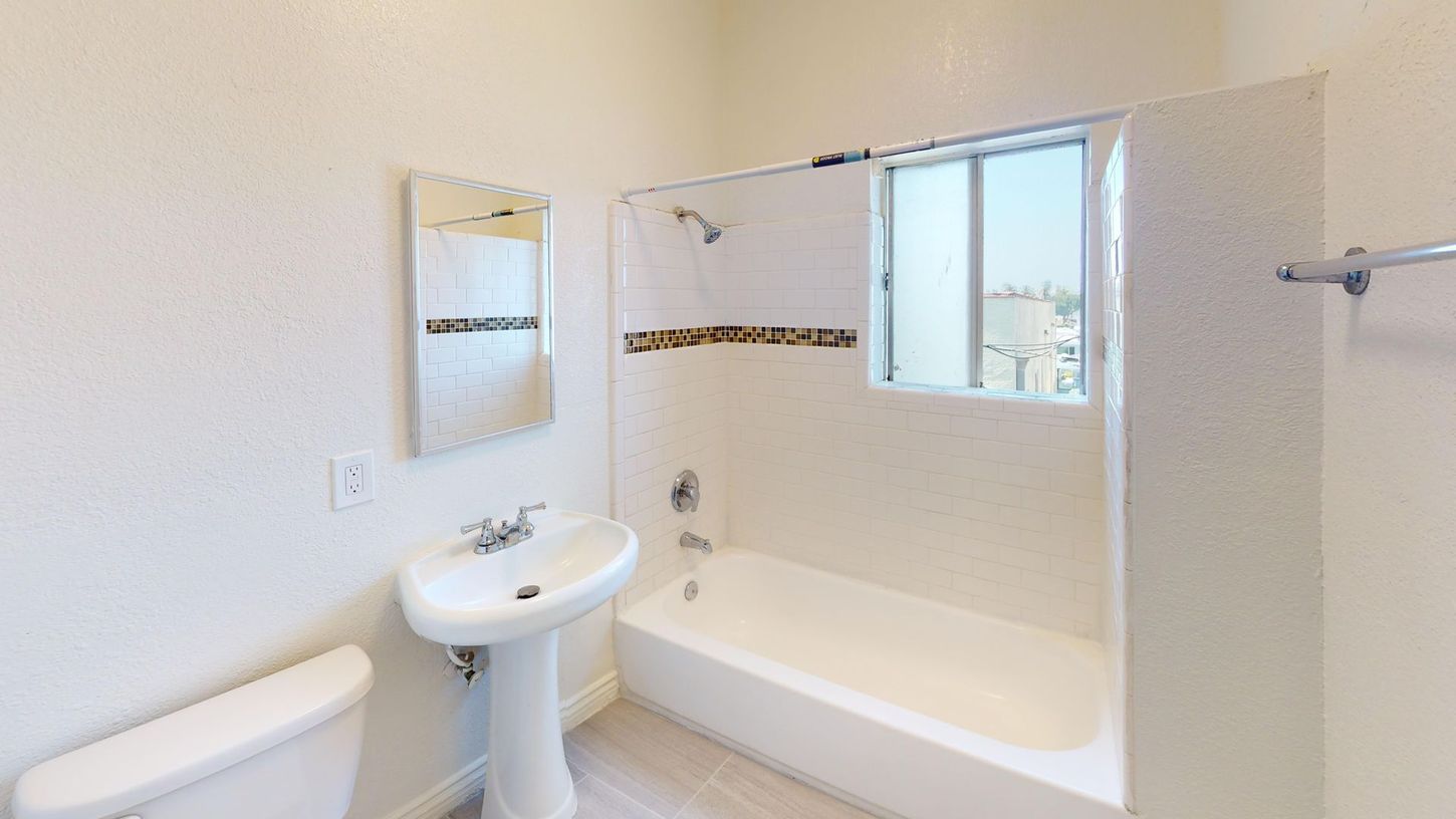 Bathroom with white fixtures: toilet, pedestal sink, bathtub/shower, mirror, window, and towel rack.