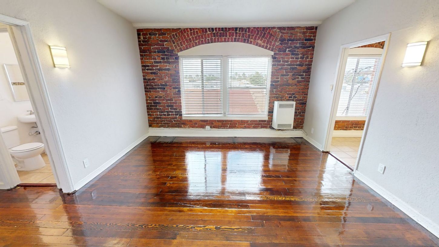 Empty apartment interior with exposed brick wall, large window, and hardwood floors.
