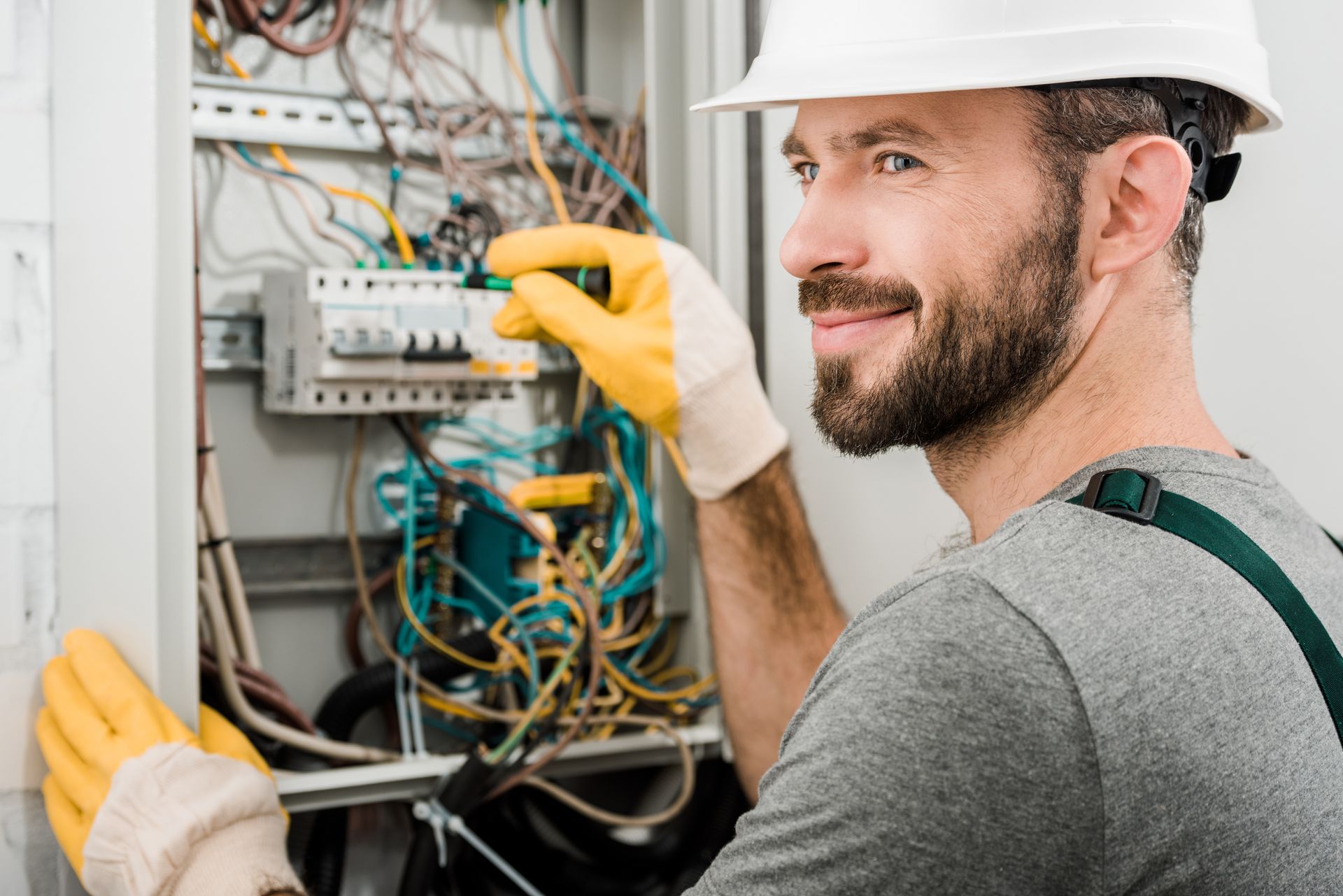 A smiling electrician expertly repairs an electrical box in a well-lit corridor, confidently using a screwdriver to complete the task.