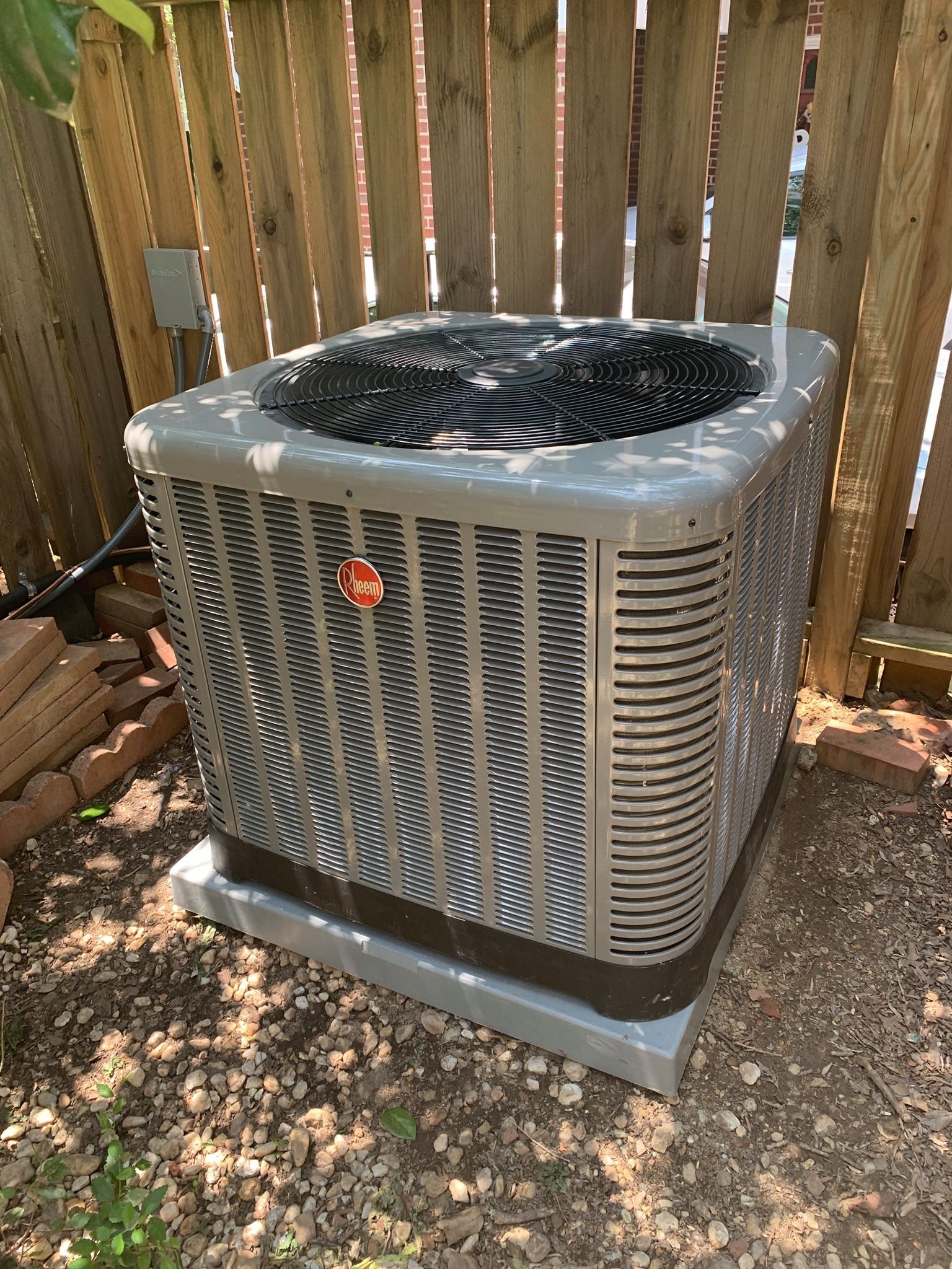 Gray Rheem air conditioner unit outside, next to a wooden fence.