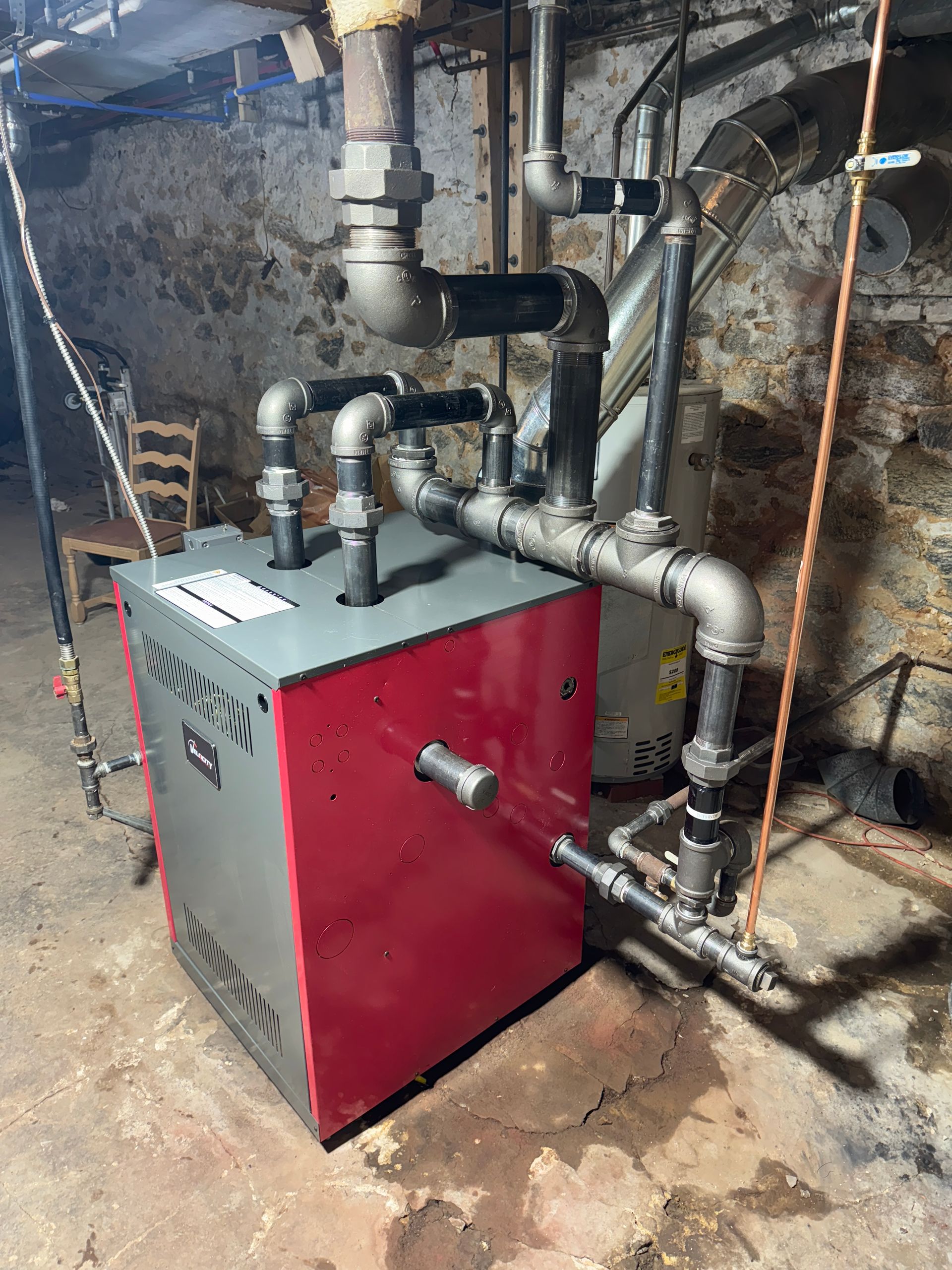 Red and gray furnace with black pipes in a basement against a stone wall.