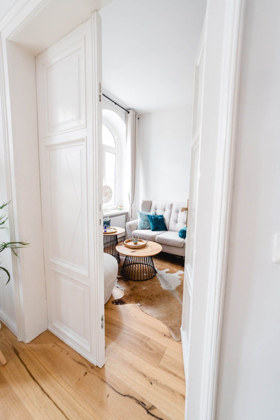 Durch eine weiße Tür gelangt man in ein helles Wohnzimmer mit Holzboden und einer Couch.