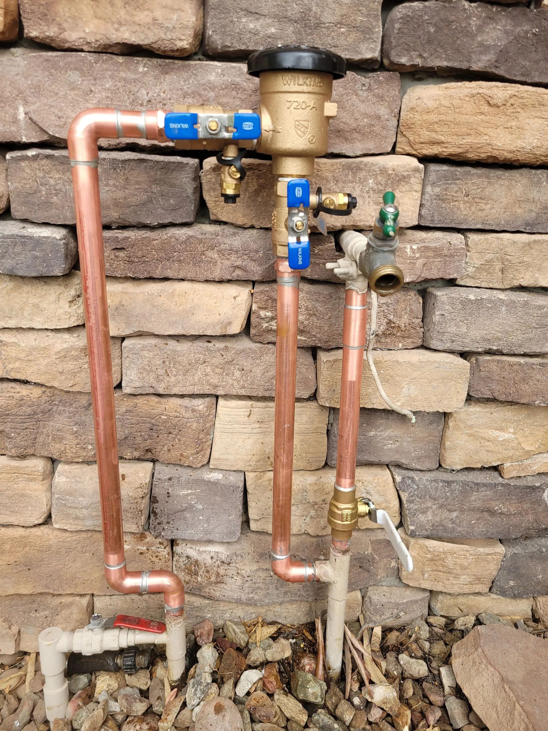 Copper plumbing with valves and water meter against a stone wall.