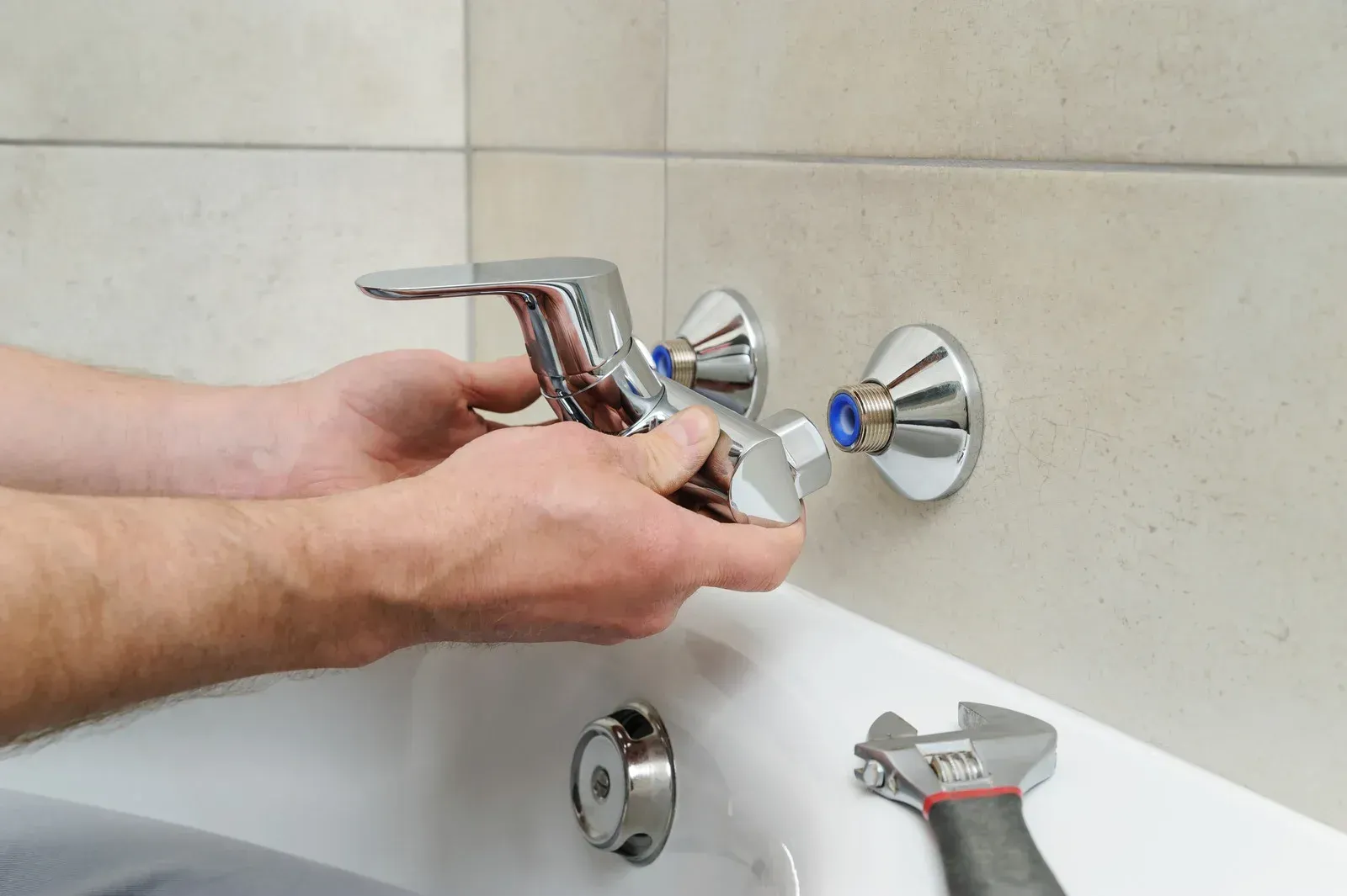 Hands installing a chrome faucet in a bathtub, with a wrench visible nearby.