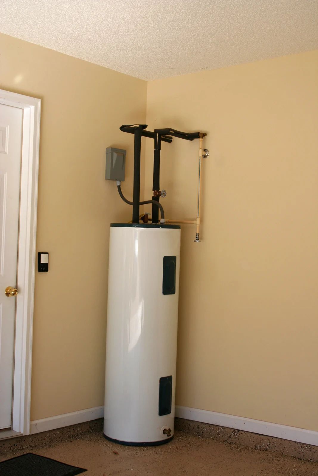 White water heater in a corner against beige walls, near a door and electrical box.