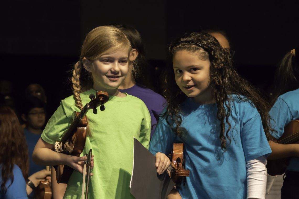 Two children smiling holding violins at CMSS concert, with background of other children.