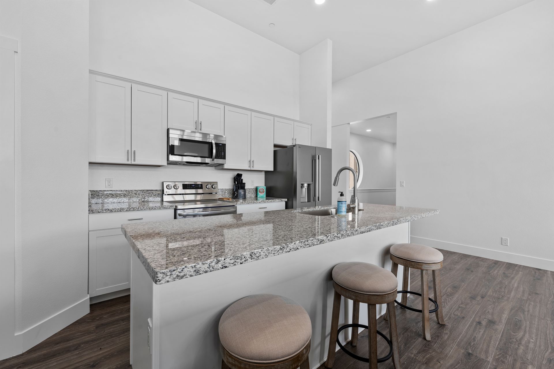 A kitchen with white cabinets , granite counter tops , stools and a refrigerator.