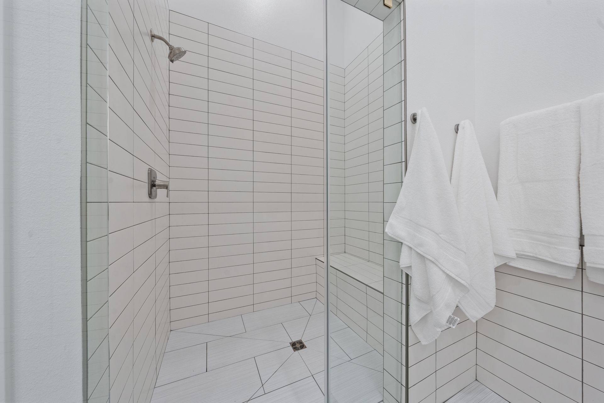 A bathroom with white tiles and a walk in shower with a glass door.