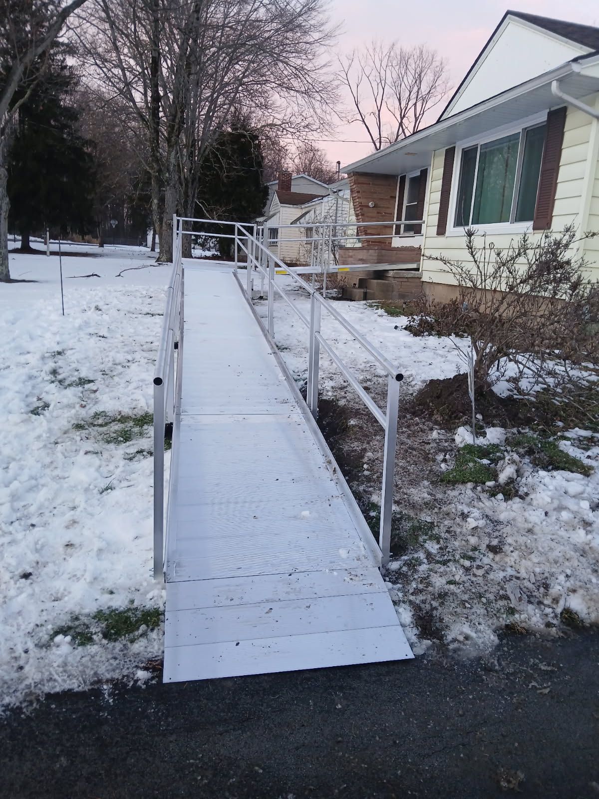 Aluminum ramp covered in snow leads to a house with a light pink sky in the background.