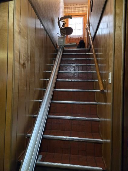 Stairlift on a narrow, brown-paneled staircase with reddish tile steps. Sunlight shines from a window.