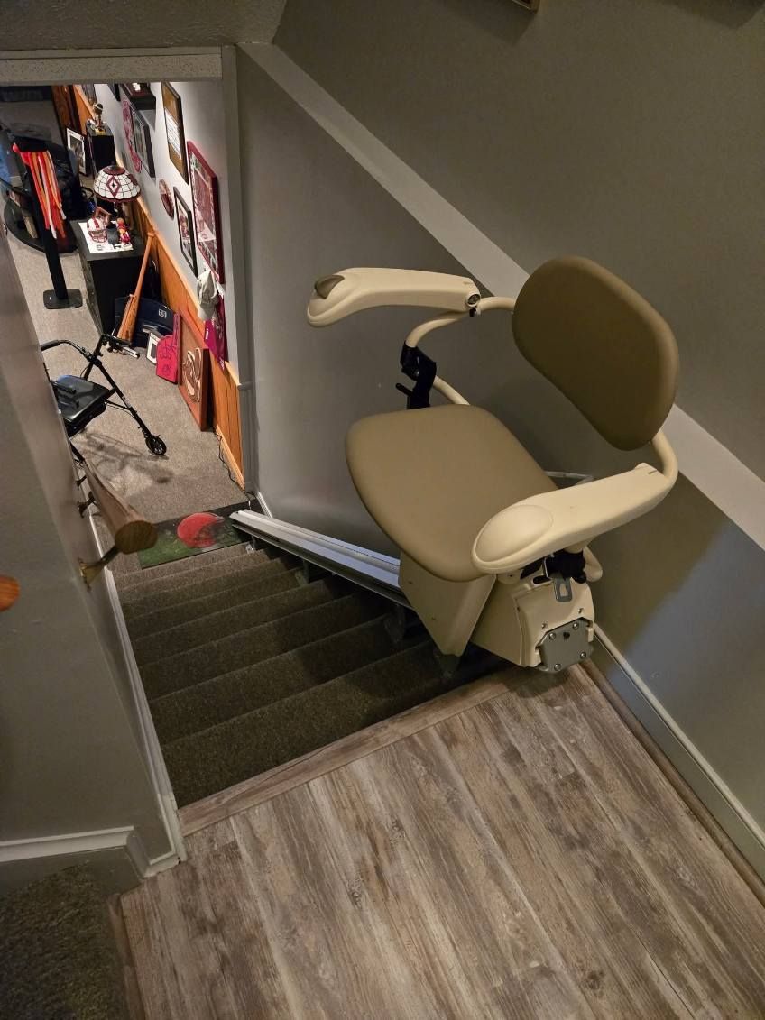Stair lift on a wooden staircase with a tan seat and armrests, leading down to a basement area.