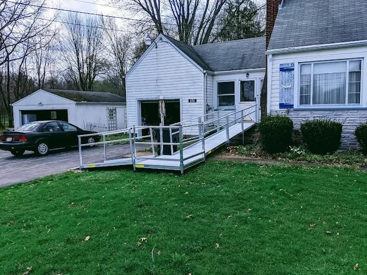 Wheelchair ramp leading to a small white house. A black car is parked next to a garage. Green grass in foreground.