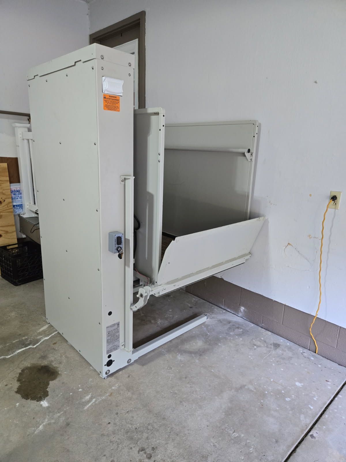 An off-white vertical platform lift installed in a garage, with the platform door open.