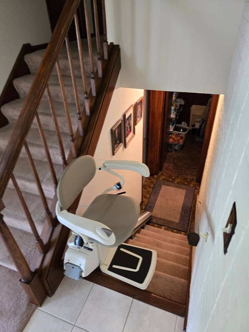 Stairlift on carpeted stairs. Wooden banister and steps. Beige lift chair. Leads to a hallway.