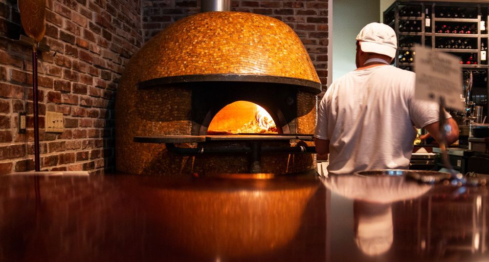 Pizza Maker Placing Pizza in a Wood-fired Oven With Exposed Brick Walls — Chefs2Go in Ayr, QLD