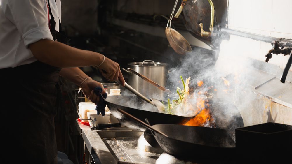 Chef Cooking With Wok, Flames Rising, Steam, and Metal Utensils in a Restaurant Kitchen — Chefs2Go in Mackay, QLD