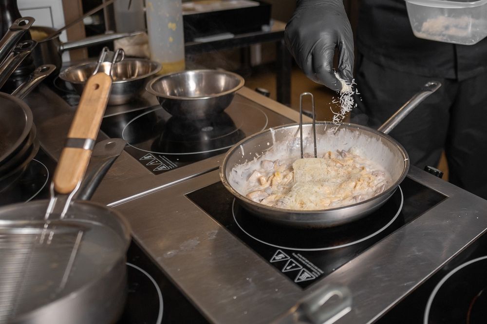 Chef in Black Glove Sprinkles Grated Cheese Into a Pan on a Stovetop — Chefs2Go in Mackay, QLD