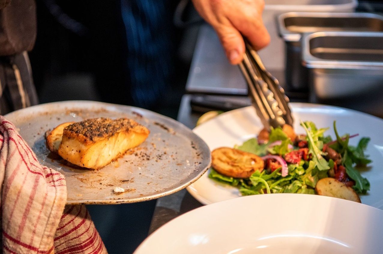 A Chef Plates Food, Placing a Seared Fish Fillet Next to a Salad on a White Plate — Chefs2Go in Bundaberg, QLD
