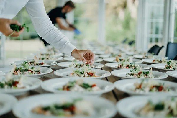 Person Garnishing Plates of Food on a Long Table in a Bright Room — Chefs2Go in Woodwark, QLD
