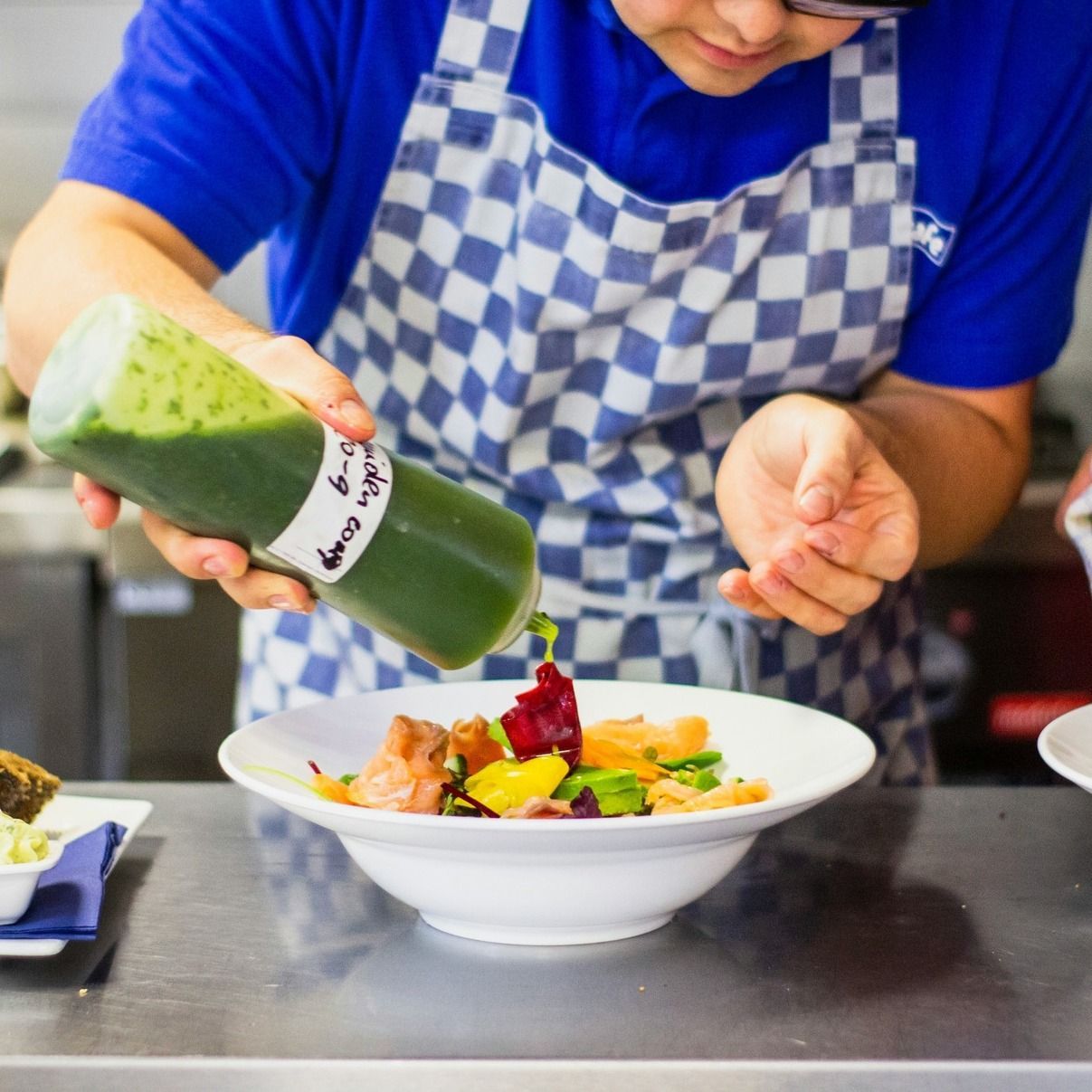 Chef in Blue Pours Green Sauce Over Salad in a White Bowl — Chefs2Go in Rockhampton, QLD