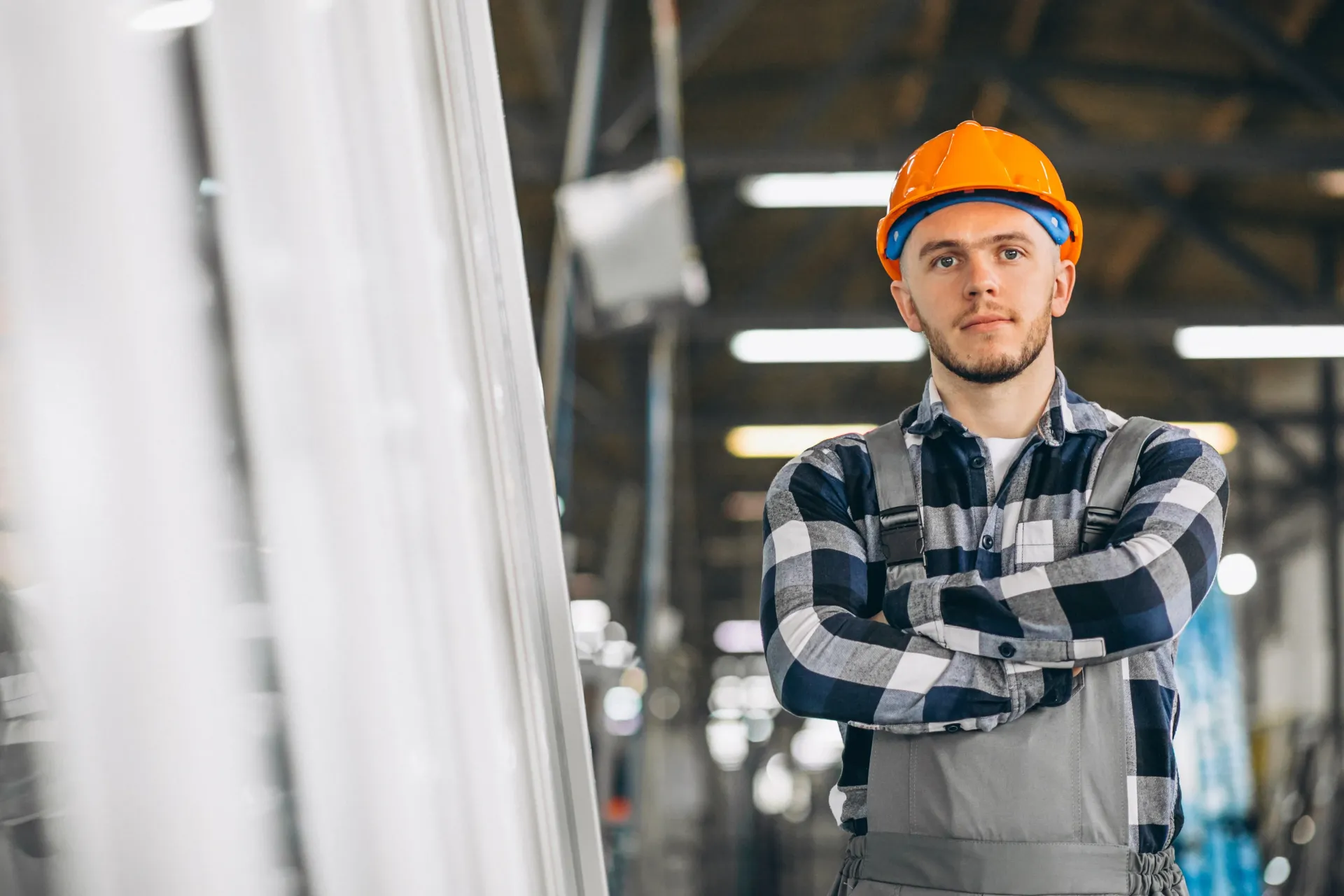 Un homme portant un casque et une salopette se tient debout dans une usine, les bras croisés.