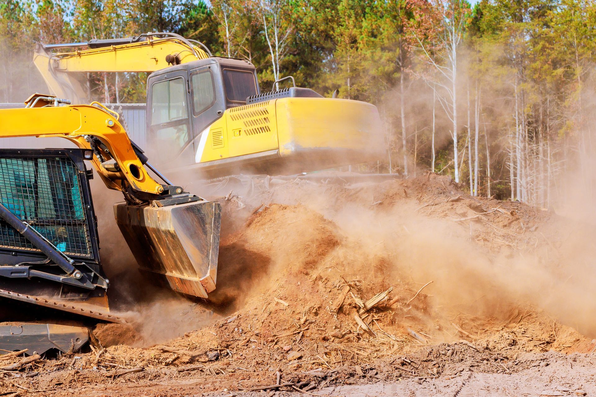 Two yellow excavators digging into a pile of dirt, kicking up dust; trees in background.