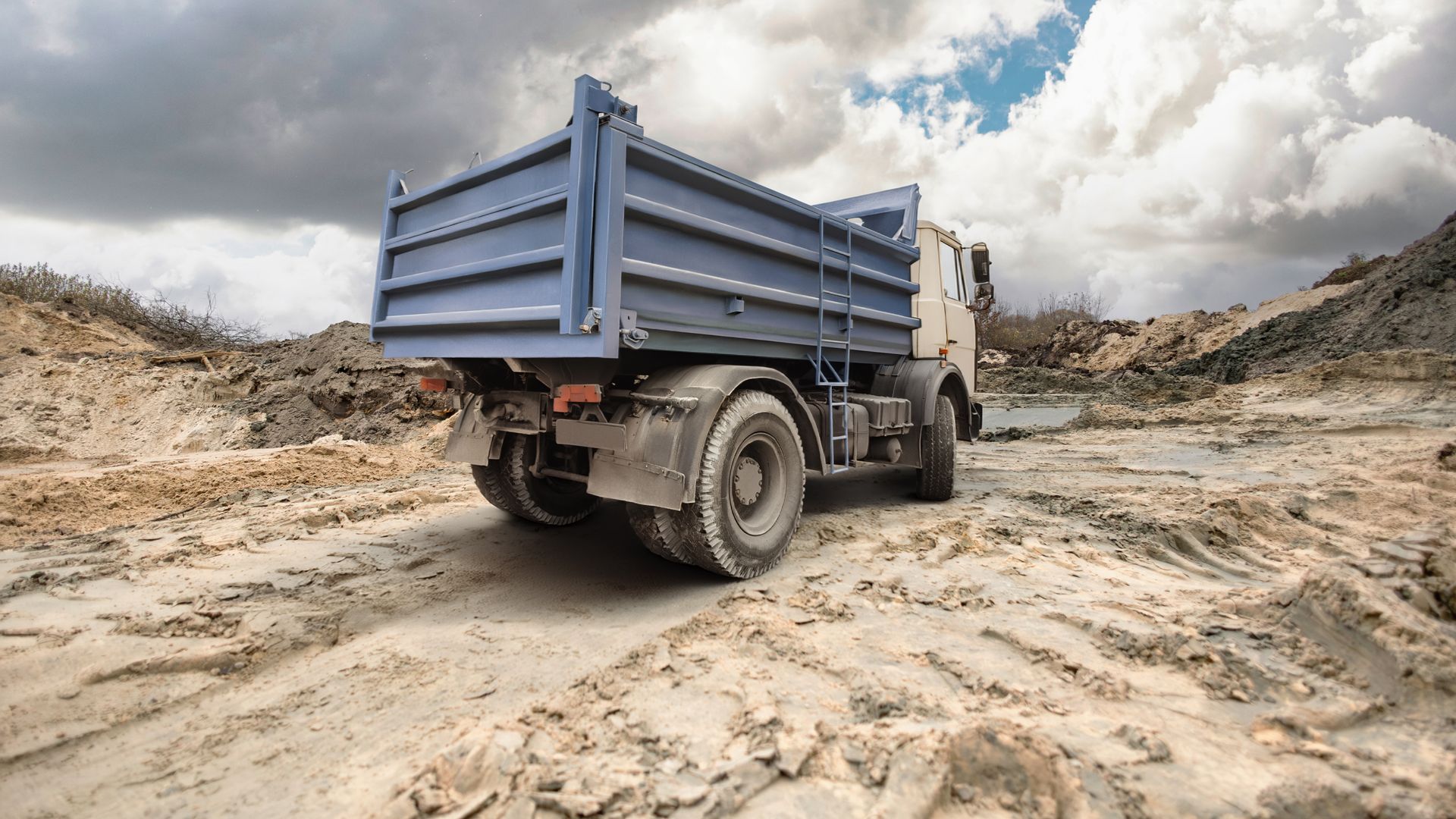 Blue dump truck on a muddy dirt road, under a cloudy sky.