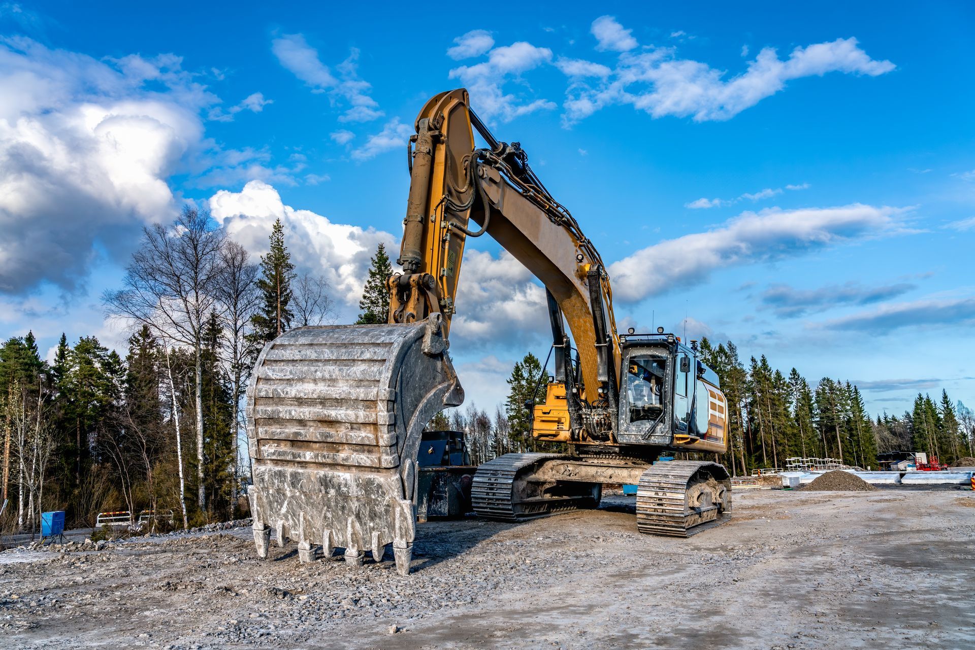 Yellow excavator on a gravel lot under a partly cloudy blue sky. Trees in the background.