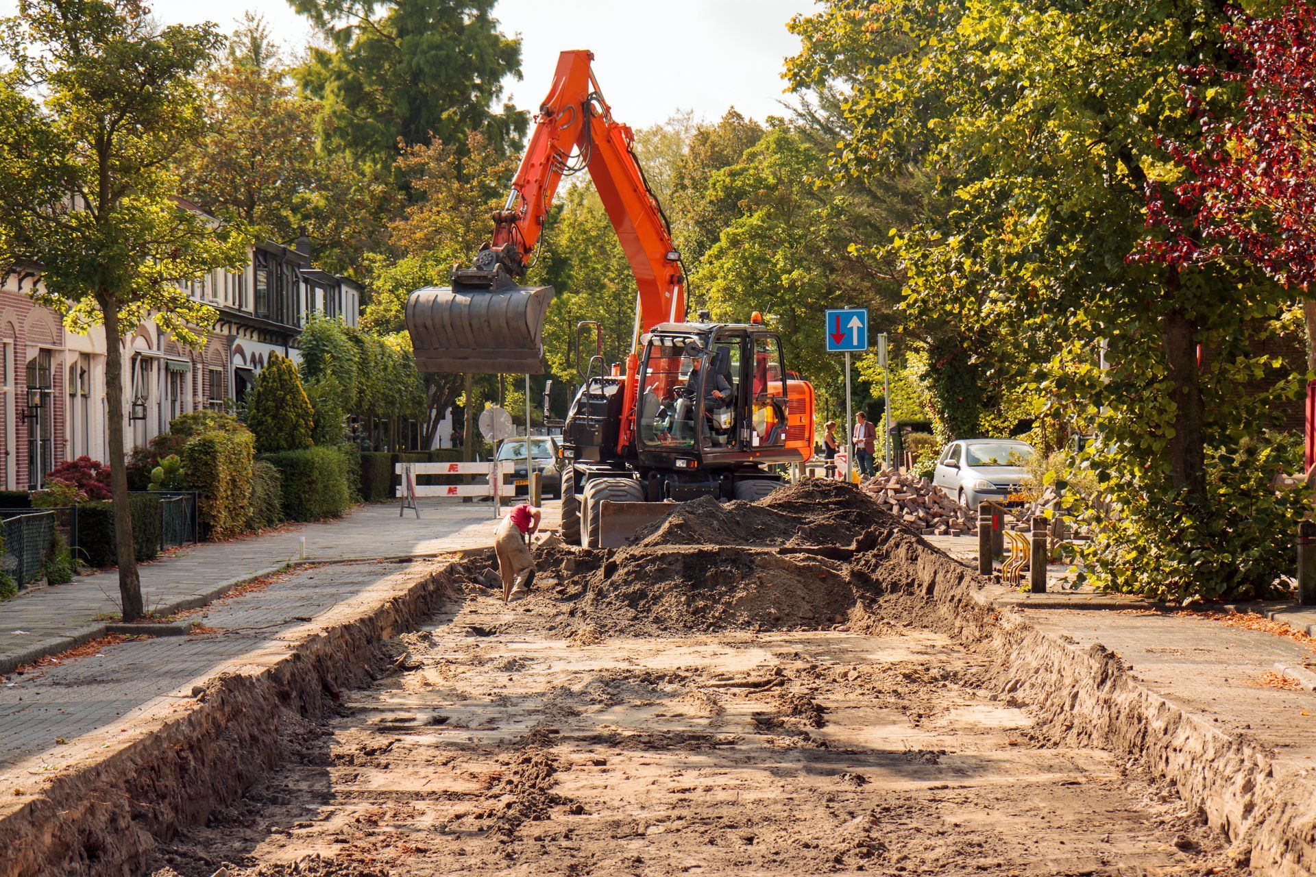 An orange excavator digging up a trench on a tree-lined street with houses.