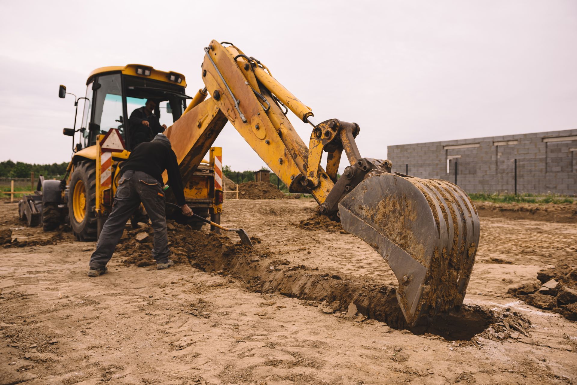 A person operating a yellow backhoe digging a trench in dirt at a construction site.