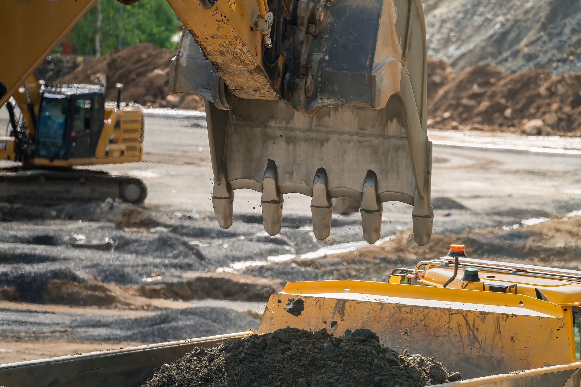 Excavator bucket dumping dirt into a yellow dump truck at a construction site.