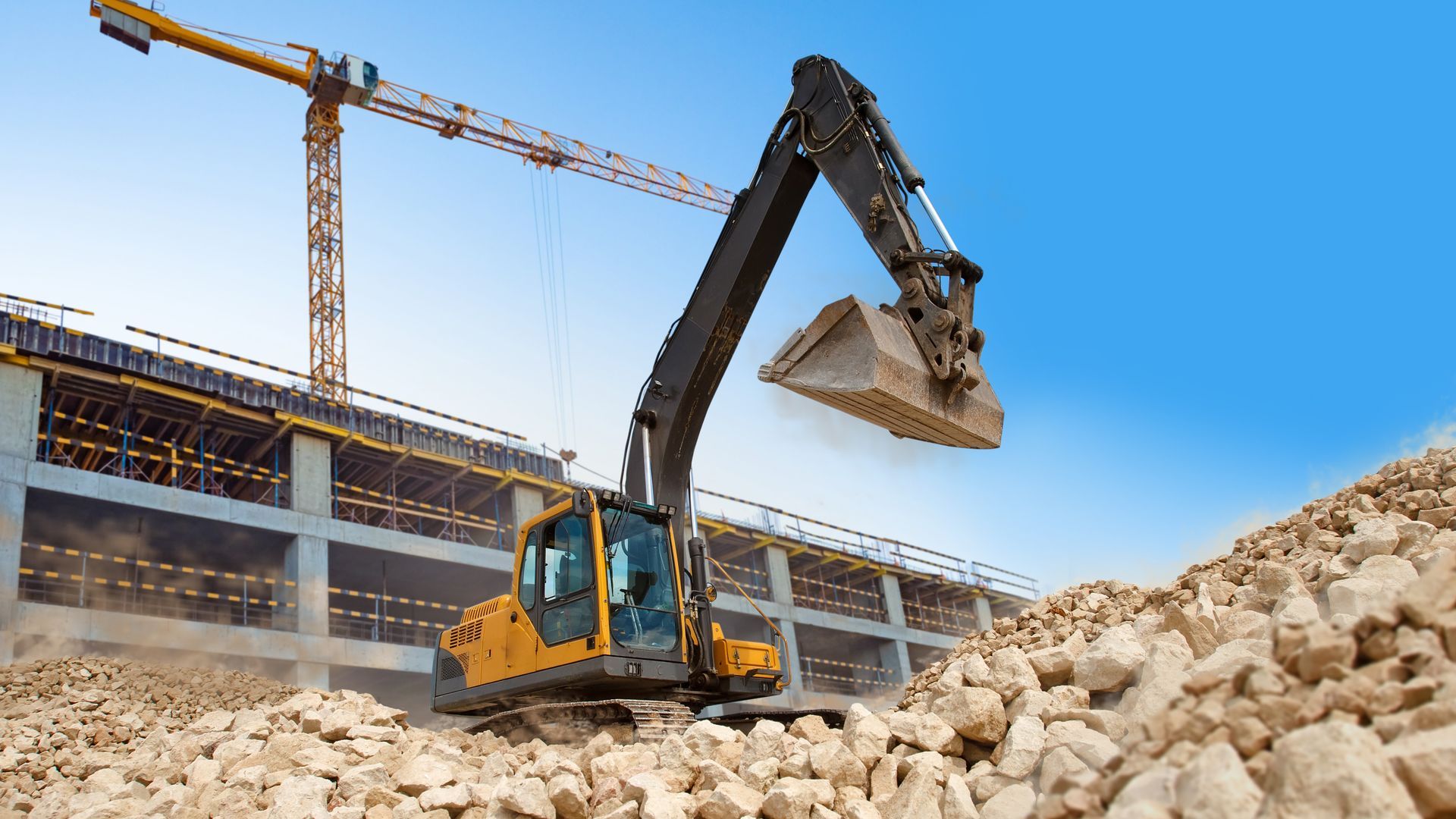 Yellow excavator moving rocks on a construction site with a crane and building in the background.