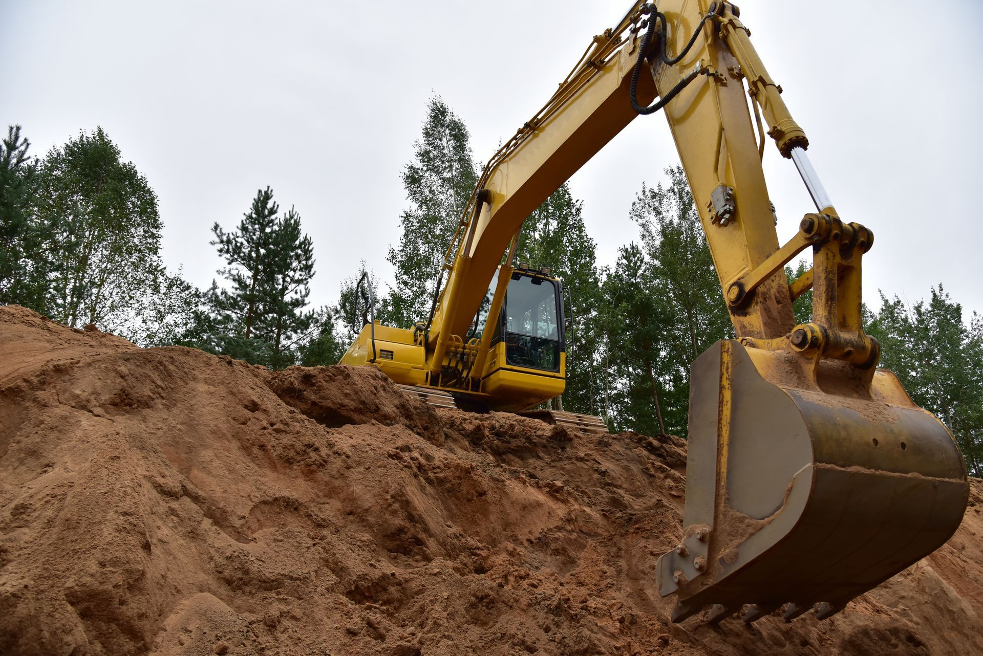 Yellow excavator digging into a mound of brown soil, trees in the background under an overcast sky.