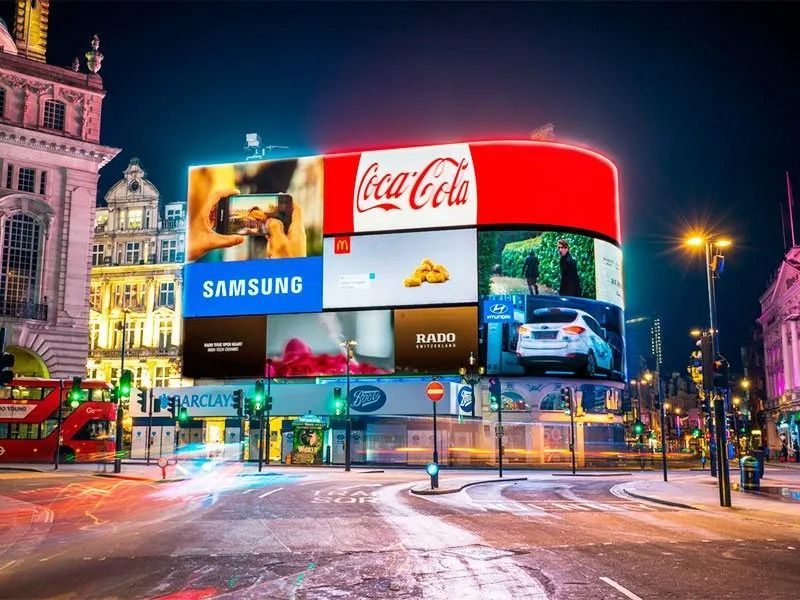 Plaza de la ciudad nevada por la noche, con brillantes vallas publicitarias digitales y autobuses rojos de dos pisos.