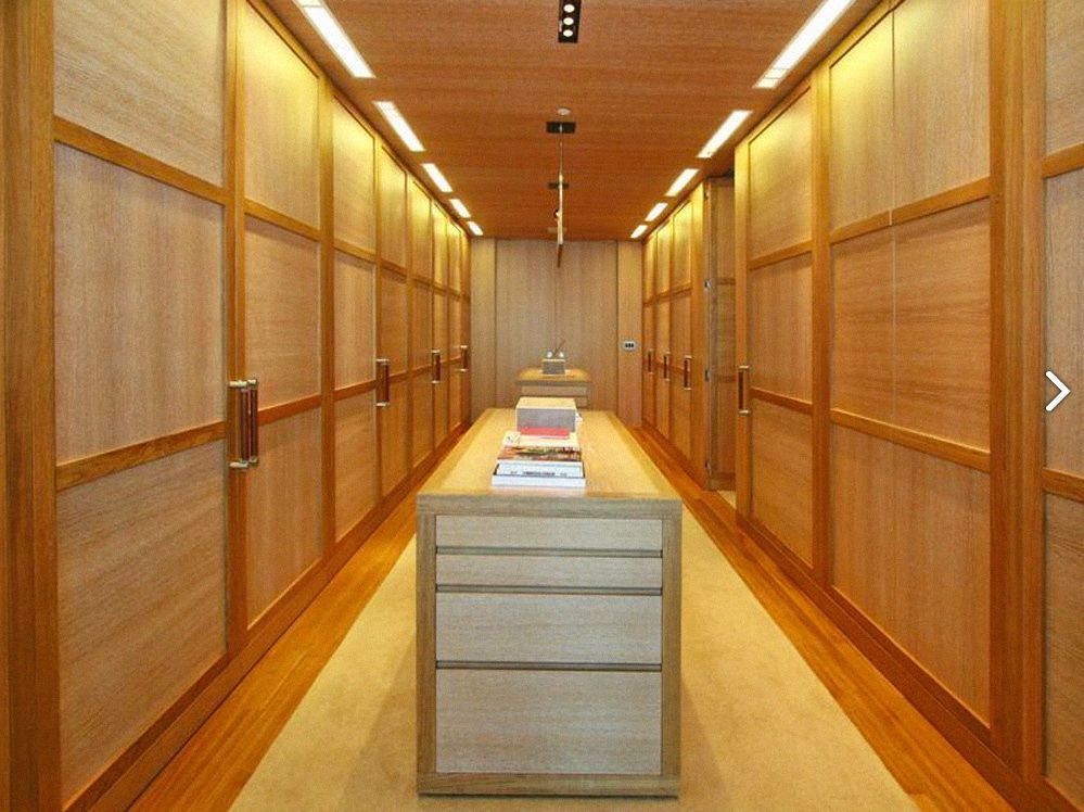 Long wooden walk-in closet with central storage island. Neutral tones, well-lit, minimalist design.