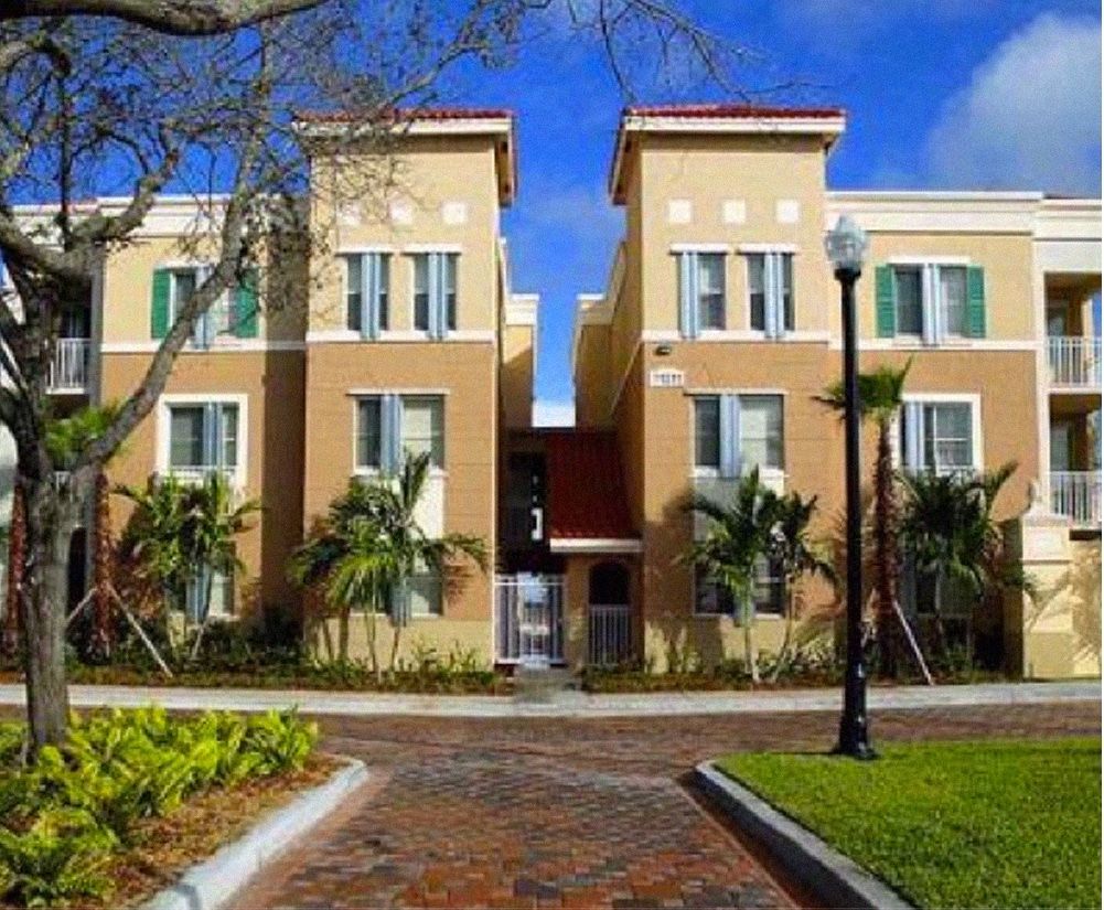 Apartment building with tan walls, green shutters, palm trees, and a brick walkway.